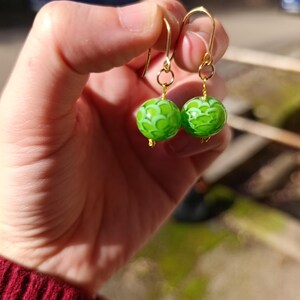 May include: A pair of handmade earrings featuring bright green, patterned glass beads. The beads are round and have a delicate, swirling design in a lighter shade of green. The earrings have gold-colored ear wires.