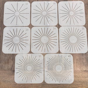 Op de afbeelding: Een set van zeven vierkante sjablonen met een sunburst-ontwerp. Elke sjabloon heeft een verschillend aantal stralen, variërend van 12 tot 48. De sjablonen zijn gemaakt van een lichtgekleurd materiaal en liggen op een houten oppervlak.