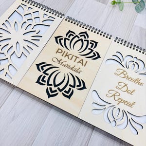 May include: A wooden spiral-bound journal with laser-cut designs and the words "PIKITAI Mandala" and "Breathe Dot Repeat." The journal features a light wood colour with black and gold lettering and floral designs.