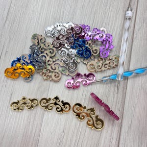 Puede incluir: Una colección de adornos decorativos de acrílico en varios colores, incluyendo dorado, plateado, azul, morado y rosa. Los adornos presentan un diseño ornamental y arremolinado. También hay dos herramientas de nail art.