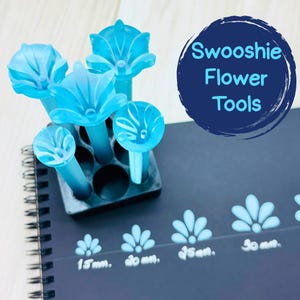 May include: A collection of light blue flower-shaped tools with stems, displayed in a black holder. The tools are next to a dark blue surface with the text "Swooshie Flower Tools" and flower impressions with size measurements.