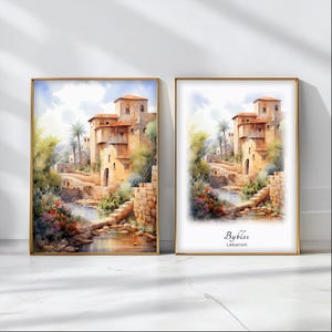 May include: Two watercolour paintings of a small village in the Middle East. The paintings are framed in gold and hung on a white wall. The painting on the left shows a view of the village from a distance, while the painting on the right shows a closer view of the village. The painting on the right also includes the text "Byblos, Lebanon".