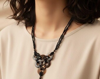 May include: A black beaded necklace with a pendant featuring interlocking brown and black rings. The necklace has a tassel of black beads at the bottom and is worn against a neutral-colored top.