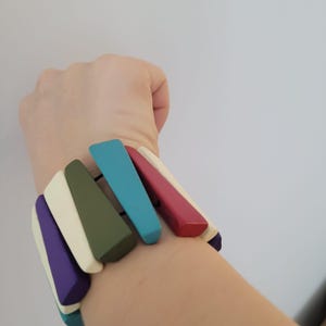 May include: A multi-coloured wooden bracelet featuring rectangular beads in shades of purple, white, green, blue, and red. The beads are connected by black elastic. The bracelet is worn on a wrist.