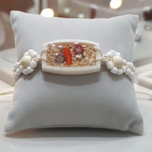 May include: A handmade bracelet featuring a rectangular, clear resin centerpiece with a red pepper, small stones, and flowers. The bracelet has white beaded flower accents and a woven cord. The bracelet is displayed on a gray pillow.