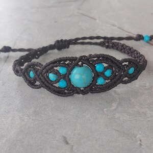 May include: A dark brown macrame bracelet featuring a central round turquoise bead surrounded by smaller turquoise beads. The intricate knotted cord design creates a symmetrical pattern, and the bracelet includes adjustable sliding knots for closure.