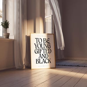 May include: A framed print with the text "TO BE YOUNG GIFTED AND BLACK" in black lettering on a white background. The frame is gold and the print is hanging on a wall in a room with white curtains.