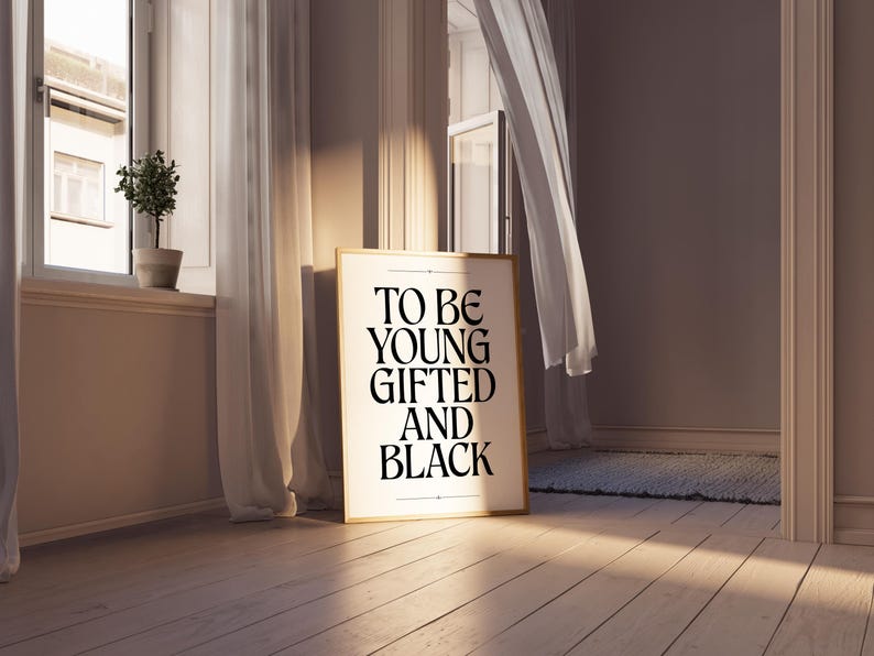 May include: A framed print with the text "TO BE YOUNG GIFTED AND BLACK" in black lettering on a white background. The frame is gold and the print is hanging on a wall in a room with white curtains.