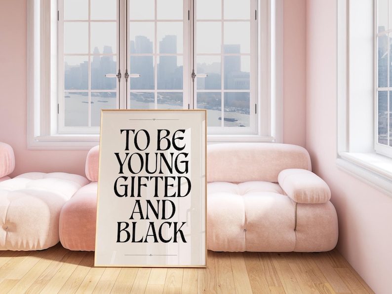 May include: A framed print with the text "To be young gifted and black" in black lettering on a white background. The print is hanging on a pink wall above a pink sofa.