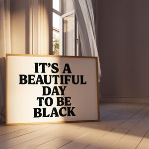 May include: A framed art print with the text "IT'S A BEAUTIFUL DAY TO BE BLACK" in bold black letters. The artwork has a light-colored background and a wooden frame. The print is displayed on a wooden floor.