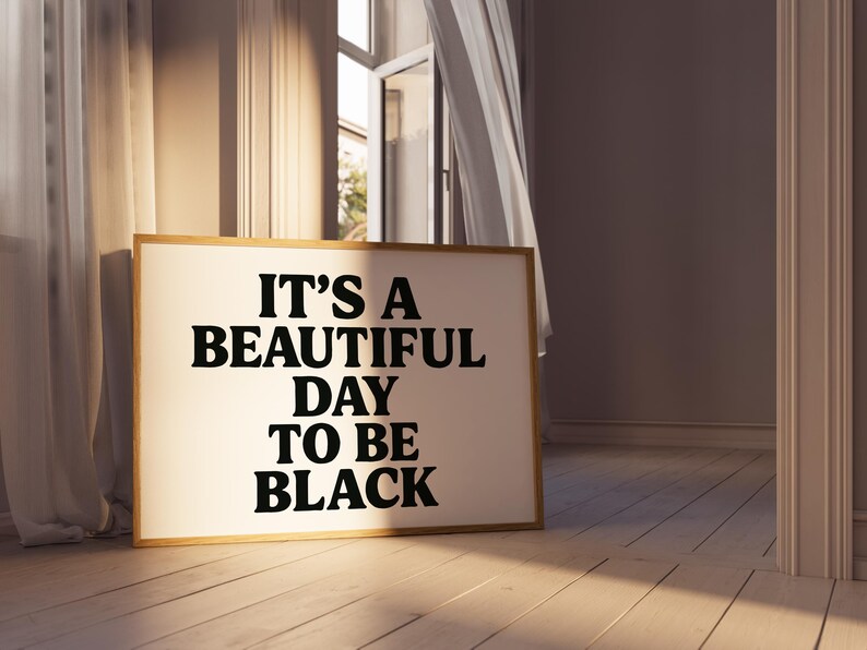 May include: A framed art print with the text "IT'S A BEAUTIFUL DAY TO BE BLACK" in bold black letters. The artwork has a light-colored background and a wooden frame. The print is displayed on a wooden floor.