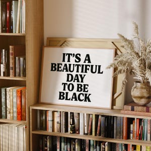 May include: A framed print with the text "IT'S A BEAUTIFUL DAY TO BE BLACK" in bold black letters. The print is displayed on a wooden bookshelf, surrounded by books and decorative items.