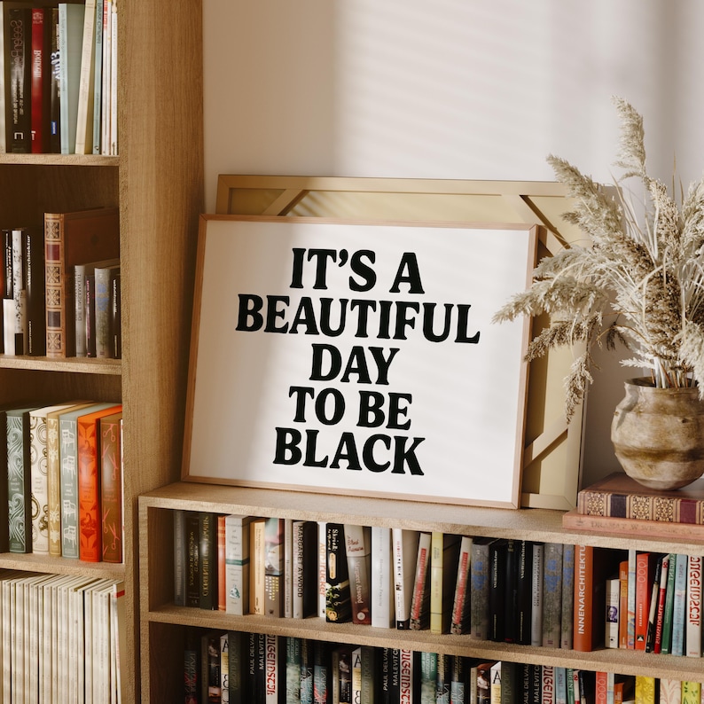 May include: A framed print with the text "IT'S A BEAUTIFUL DAY TO BE BLACK" in bold black letters. The print is displayed on a wooden bookshelf, surrounded by books and decorative items.