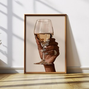 May include: A hand with jeweled nails holds a glass of white wine. The glass is clear and has a long stem. The hand is brown and has a gold ring on one finger.