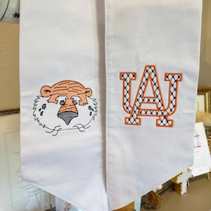 May include: White graduation stole with embroidered tiger face and the letter "A" with a blue and orange pattern. The stole is made of a soft fabric and is designed for graduation ceremonies.
