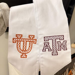 May include: Two white graduation stoles with embroidered university logos. One stole features an orange "UT" logo, and the other displays a maroon "A&M" logo. The stoles are made of a smooth, white fabric.