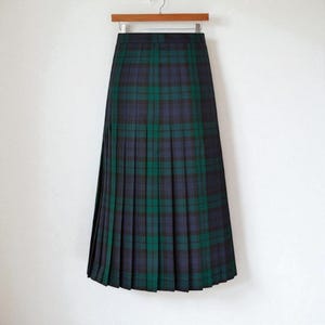 May include: A long, pleated tartan skirt in shades of dark green, navy blue, and black, displayed on a wooden hanger. The skirt's full length is visible, highlighting the intricate pleating and the traditional check pattern against a white backdrop.
