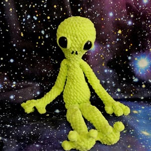 May include: A lime green crocheted alien doll with large black eyes, long arms, and three-fingered hands and feet. The alien is sitting against a starry background, giving it a space-themed appearance. The doll is made of a soft, textured yarn.