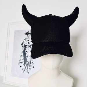 Puede incluir: Una gorra de béisbol negra con dos cuernos negros en la parte superior. La gorra está en una cabeza de maniquí blanca.