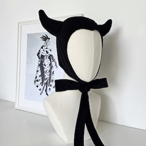 May include: Black velvet devil horns hat with a long black ribbon tie. The hat is on a white mannequin head.