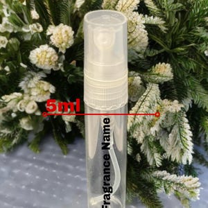 May include: A clear 5ml fragrance sample bottle with a white sprayer cap. The bottle has the words "Fragrance Name" printed vertically. The image shows the bottle against a backdrop of green and white foliage.
