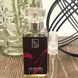 May include: A clear perfume bottle with a silver cap and a smaller clear spray bottle. The perfume bottle has a dark label with the text "FING UNBELIEVABLE" and a graphic. The bottles are on a wooden surface.