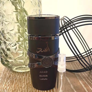 May include: A black perfume bottle with silver accents and the text "ASAD ELIXIR Lattafa". The bottle has a cylindrical shape with a decorative band. A small, clear spray bottle is next to it.