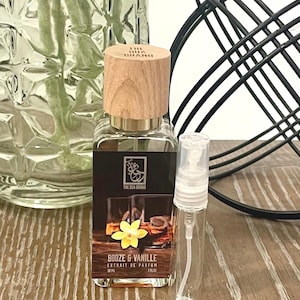 May include: A perfume bottle with a wooden cap and a clear glass body. The label reads "Booze & Vanille Extrait de Parfum" with a picture of a drink and a yellow flower. A small, clear spray bottle is next to it.