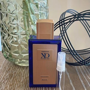 May include: A blue perfume bottle with a gold cap and a small, clear sample vial. The bottle has a gold label with the text "ORIENTICA XO XCLUSIF OUD BLEU". A clear glass vase with green stems is in the background.