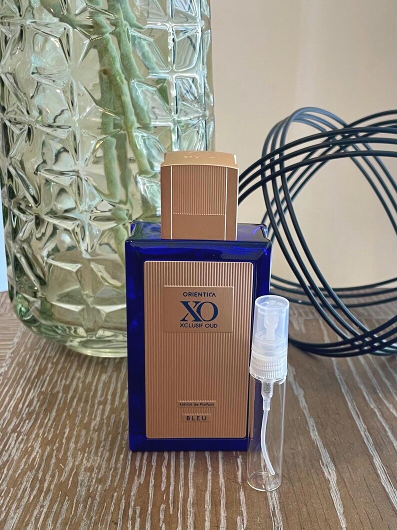 May include: A blue perfume bottle with a gold cap and a small, clear sample vial. The bottle has a gold label with the text "ORIENTICA XO XCLUSIF OUD BLEU". A clear glass vase with green stems is in the background.