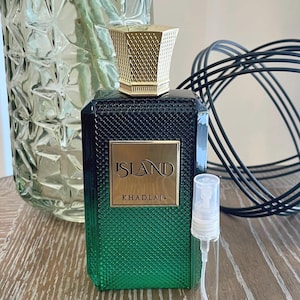 May include: A perfume bottle with a gold cap and a gradient green and black body. The bottle has a gold label that reads "ISLAND KHADLAJ." A small, clear sample vial is next to the bottle. The background includes a glass vase and black decorative wire.