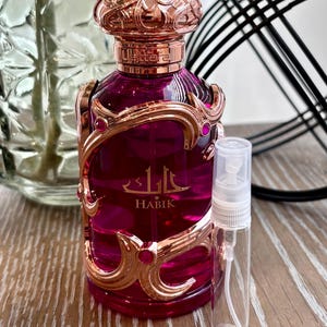 May include: A magenta perfume bottle with ornate rose gold accents and a matching cap. The bottle has the word "HABIK" in gold lettering. A small, clear spray bottle is next to the perfume bottle.