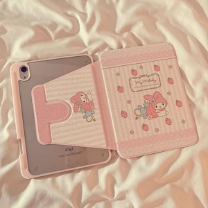 May include: A pink iPad case featuring a My Melody design. The case has a pink and white striped pattern with strawberries and flowers. The case is open, revealing the iPad.