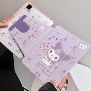 Peut inclure: Une housse de tablette lilas avec un personnage de dessin animé tenant un ballon en forme de cœur. L'étui a un rabat avec un motif répété de nœuds, de cœurs et d'autres petites illustrations. Le texte sur l'étui se lit : « I may look tough and fearless but I'm also pretty and girly ! »