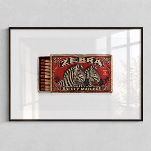 Puede incluir: Una impresión enmarcada que muestra una caja de cerillas de estilo vintage con fondo rojo y dos cebras. El texto en la caja dice ZEBRA SAFETY MATCHES y MADE IN KENYA. La caja está parcialmente abierta, revelando cerillas con puntas rojas.