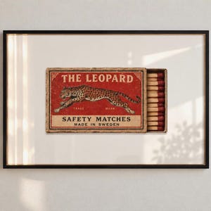 May include: A framed wall art print featuring a vintage-style matchbox labelled THE LEOPARD SAFETY MATCHES MADE IN SWEDEN. The red matchbox displays a leaping leopard illustration and contains a row of wooden matches with dark red tips.