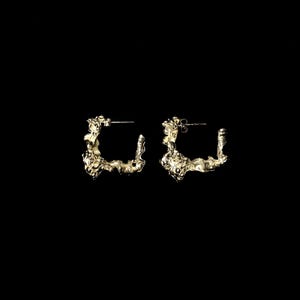 May include: A pair of gold-toned hoop earrings with a textured, organic design. The earrings are made of metal and have a slightly irregular shape.