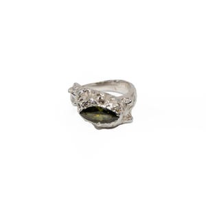May include: A silver ring with a textured, organic design. The ring features a dark green, oval-shaped gemstone at its center. The band has a rough, uneven surface, giving it a unique, handcrafted appearance. The ring is set against a white background.