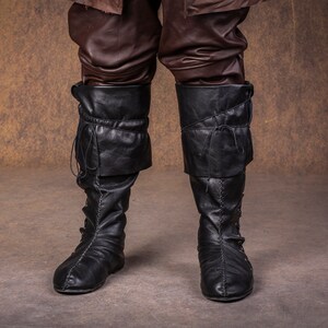 Op de afbeelding: Een paar zwarte leren laarzen met veters en gespen. De laarzen hebben een puntige neus en een lage hak.