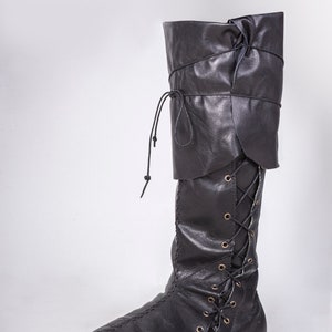 Op de afbeelding: Zwarte leren laarzen die tot aan de knie komen met een vetersluiting aan de voorkant en een omslag aan de bovenkant.