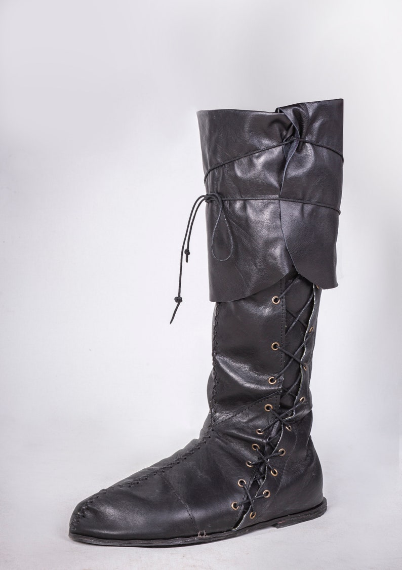 Op de afbeelding: Zwarte leren laarzen die tot aan de knie komen met een vetersluiting aan de voorkant en een omslag aan de bovenkant.