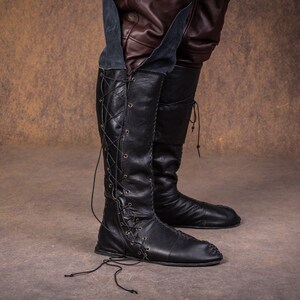 Op de afbeelding: Zwarte leren laarzen die tot aan de knie komen met veters aan de voorkant en een spitse neus.