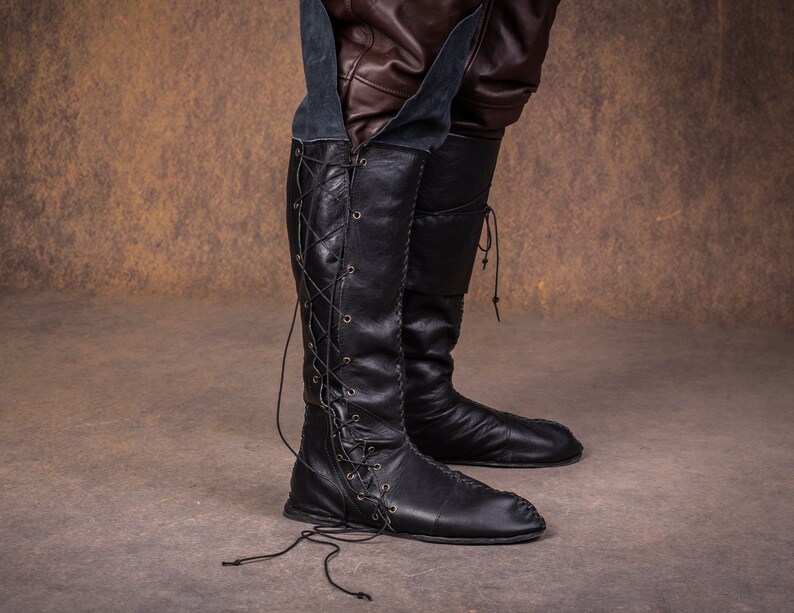 Op de afbeelding: Zwarte leren laarzen die tot aan de knie komen met veters aan de voorkant en een spitse neus.