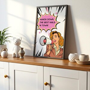 Modern shelf styling with a black framed pop art beauty poster. "Best Nails In Town" sign displayed on a white cabinet. Bold graphic design with Ben-Day dots and comic book elements. Unique gift idea for salon grand openings.