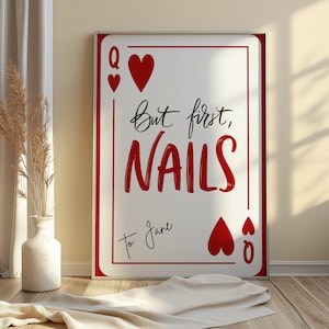 Puede incluir: Un estampado enmarcado con un diseño de carta de juego que muestra el texto "But first, NAILS" en rojo. La carta incluye corazones rojos y el texto "To Jane" en escritura negra. El marco es plateado.