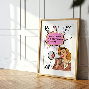 Close up view of a framed manicurist gift poster in a sunlit room. Detailed retro illustration of a woman shouting into a megaphone. High-quality printable wall art with a "Hands Down" nail pun message.