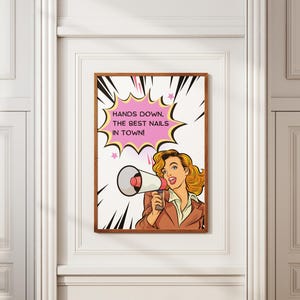 Framed "Hands Down The Best Nails In Town" pop art poster on a white wall. Retro comic book style illustration of a blonde woman holding a megaphone. Bold pink speech bubble and yellow background. Perfect marketing signage for nail salons and beauty bars.