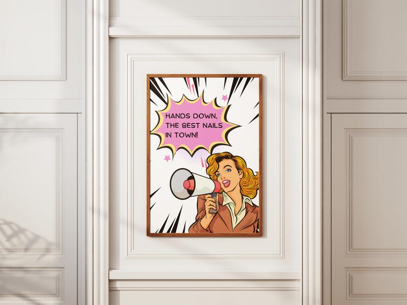 Framed "Hands Down The Best Nails In Town" pop art poster on a white wall. Retro comic book style illustration of a blonde woman holding a megaphone. Bold pink speech bubble and yellow background. Perfect marketing signage for nail salons and beauty bars.