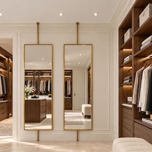 May include: Two tall, rectangular floor-to-ceiling mirrors with thin gold frames, mounted on vertical gold poles. The mirrors are positioned on a light-colored tiled floor in a walk-in closet featuring dark wood shelving units and a cream-colored bench.