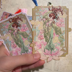May include: Bookmarks featuring a detailed illustration of a woman in a flowing green gown, surrounded by pink flowers and clouds. The bookmarks have a gold border and a tassel attached to the top.